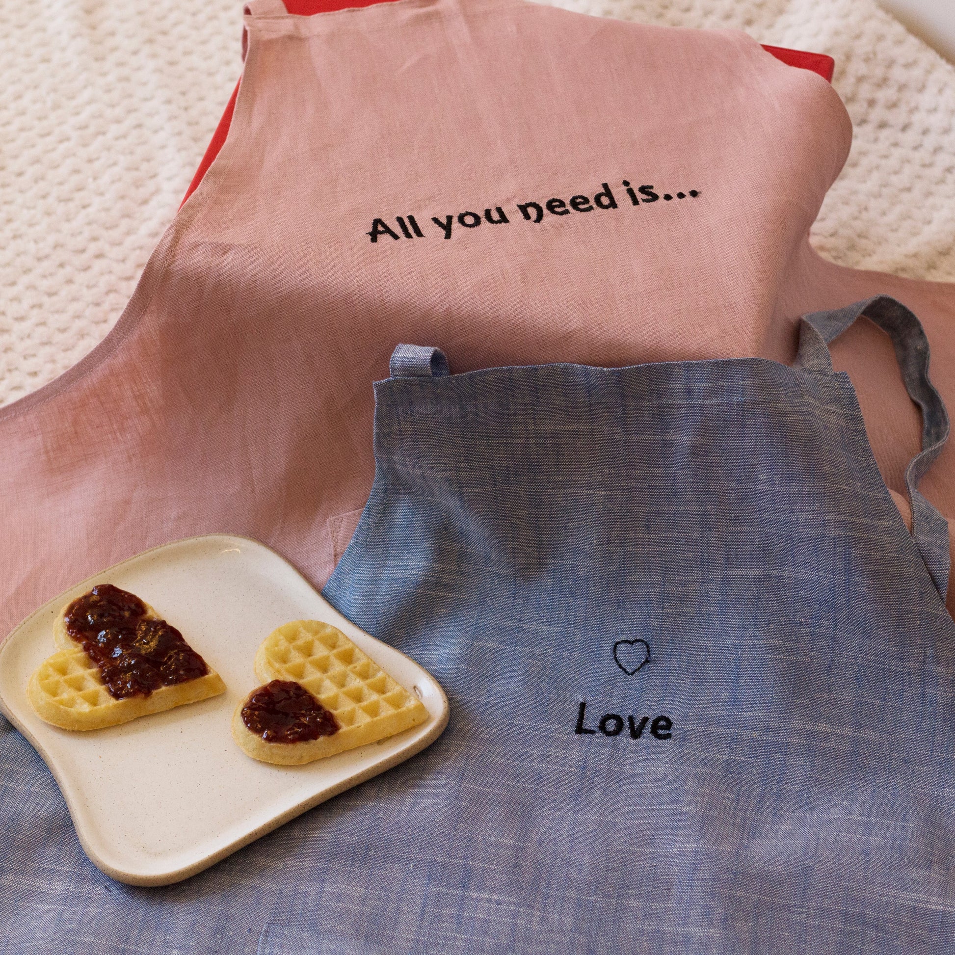 Two aprons, one pink with text and the other blue with a heart and text, on a textured surface.