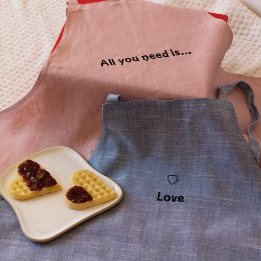 Two aprons, one pink with text and the other blue with a heart and text, on a textured surface.